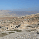 Ancient ruins on a rocky hillside with a backdrop of arid mountains under a clear blue sky.