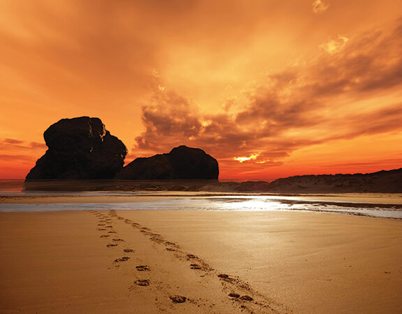Footprints on a sandy beach at sunset, with dramatic orange sky and silhouetted rock formations in the distance.