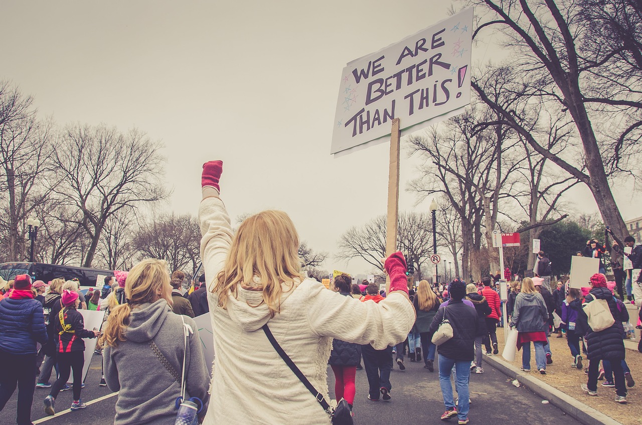 People at a protest holding signs, one says We Are Better Than This, in a city street with trees and overcast sky.