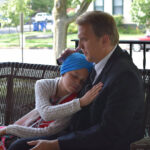 Man and child in embrace on a porch, child with oxygen tube, wearing a blue headscarf, hugging man.