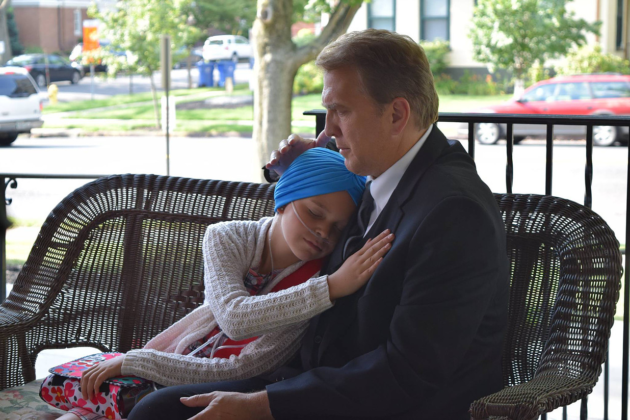 Man and child in embrace on a porch, child with oxygen tube, wearing a blue headscarf, hugging man.