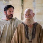 Two men in biblical costumes talking in an ancient stone setting, one with a beard and the other gesturing.