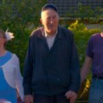 Group of three people standing outside, smiling and holding hands amidst greenery, enjoying a sunny day.