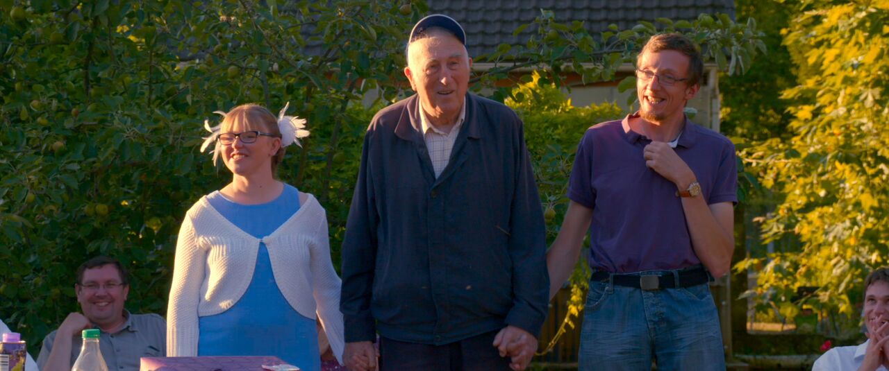 Group of three people standing outside, smiling and holding hands amidst greenery, enjoying a sunny day.