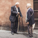 Elderly couple conversing in the street, both using canes; man in suit, woman in scarf and coat.