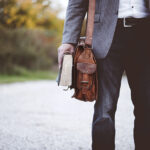 Man holding a leather bag and book, standing on a gravel path in casual attire.
