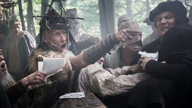 Medieval men in intense confrontation, one in fur and metal headgear, others gesturing and holding papers in a forest setting.