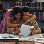 Couple reading and embracing in a library, surrounded by books and papers, with shelves in the background.