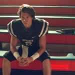 Football player in uniform sits on bleachers, appearing thoughtful.
