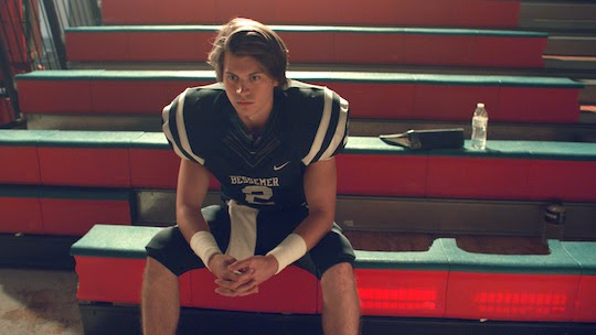 Football player in uniform sits on bleachers, appearing thoughtful.