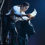 Actor reads a letter dramatically on stage with spotlight, surrounded by a ship's ropes and railings.
