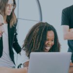 Group of diverse colleagues collaborating on laptops with smiles in a bright office space.