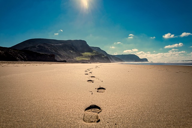 Footprints on a sunlit beach leading towards distant mountains under a clear blue sky.