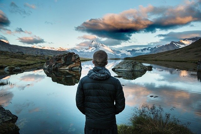 Person in a jacket stands by a reflective lake, admiring mountains during sunset. Peaceful nature scene.