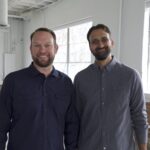 Two smiling men in a bright office setting with windows and exposed brick walls.