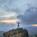 Person standing on a rock with arms outstretched, overlooking a scenic sunset with dramatic clouds.