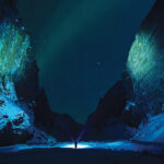 Person with flashlight in a snowy canyon under a starlit sky with green aurora borealis.