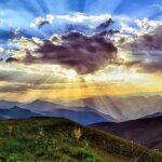 Mountain landscape at sunrise with vibrant sky, sun rays through clouds over rolling hills and wildflowers in foreground.