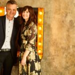 Smiling couple standing in front of rustic wall with marquee lights.