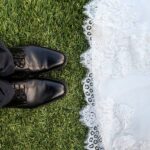 Groom's black shoes on grass next to bride's lace wedding dress hem, symbolizing marriage and new beginnings.