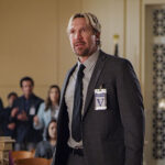 Man in a suit with visitor badge stands in a courtroom, speaking to the audience.