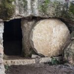 Ancient stone tomb with round entrance, partially covered by a large stone, surrounded by rocks and vegetation.
