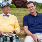 Two men sitting on a park bench, one in a colorful outfit, the other in a blue shirt, with a grassy background.