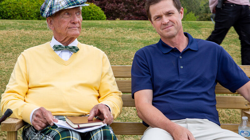 Two men sitting on a park bench, one in a colorful outfit, the other in a blue shirt, with a grassy background.