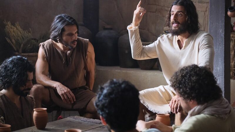 Biblical scene with a man teaching a group around a table in ancient attire, discussing with gestures and focus.