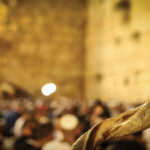Shofar horn in focus against a blurred background of a stone wall and crowd at a religious gathering.