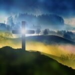 Silhouette of a cross on a misty hill with sunrise, forest, and foggy landscape in the background.