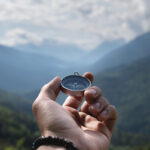 Magnetic compass in the palm of a male hand against the backdrop of a mountain range in the clouds in the summer outdoors, travel, first person view